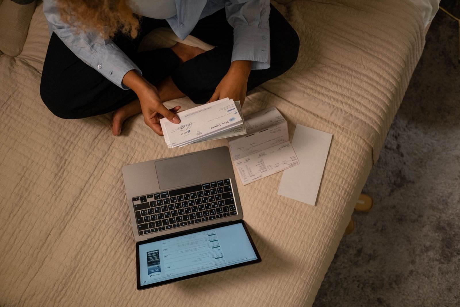 A woman looks at forms on a bed while her laptop sits open beside her