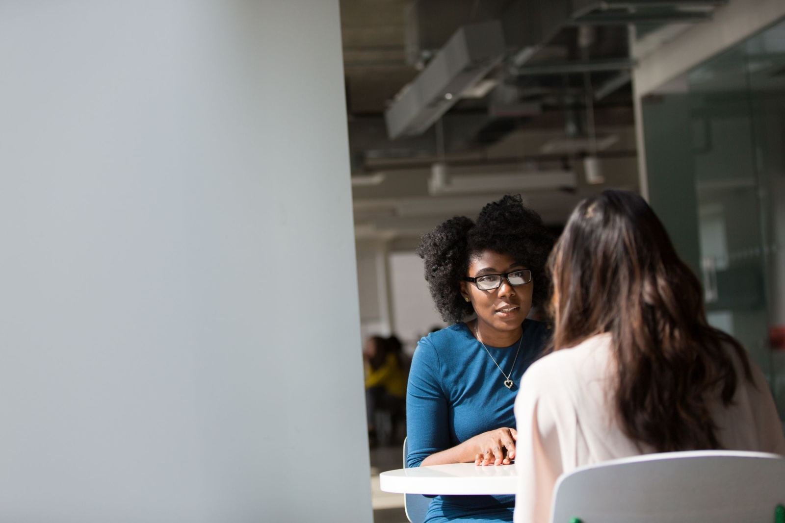 Two women sit at a white table talking in a bright office