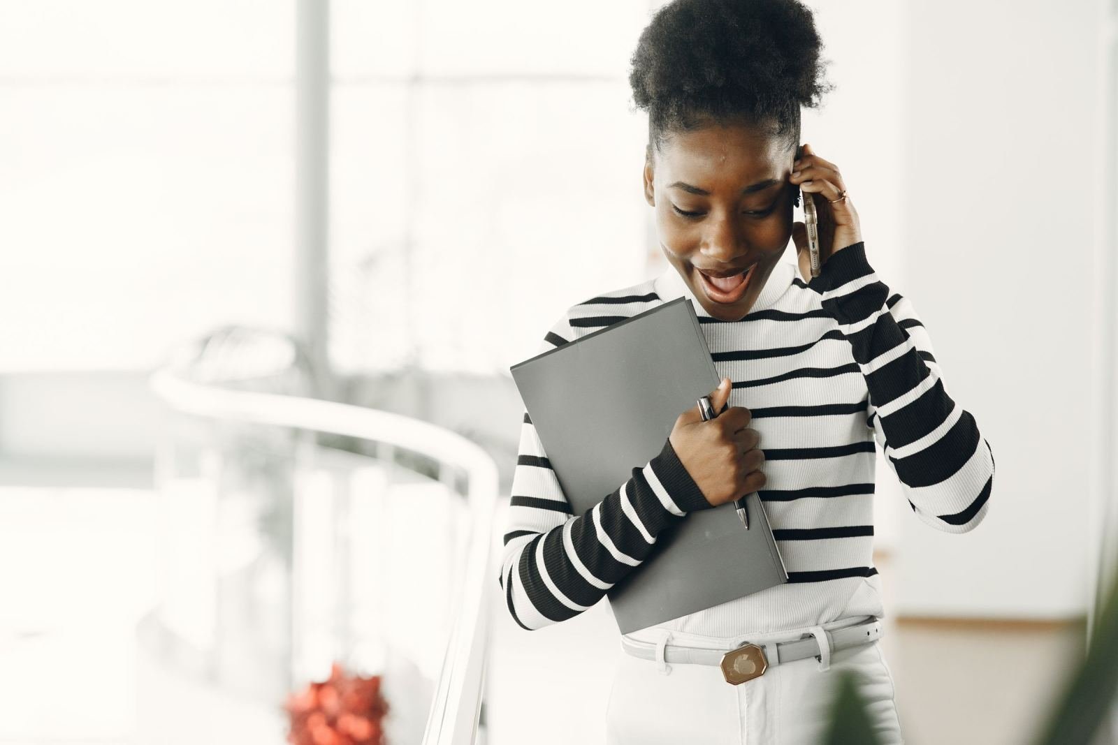 A woman in a striped top smiles and holds a folder while talking on her phone.