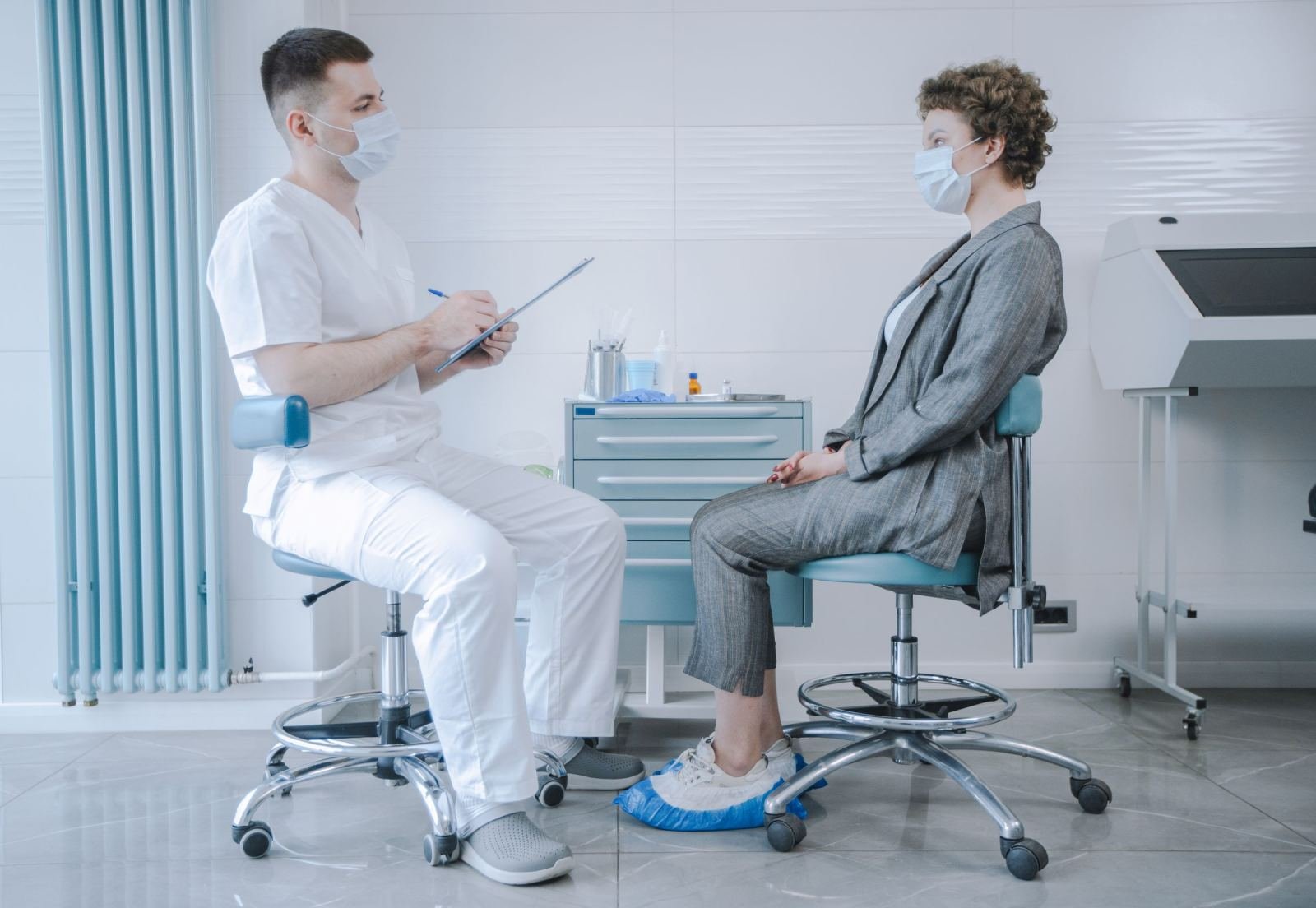 A doctor in scrubs and a patient in a suit both wearing masks talk in a clinic