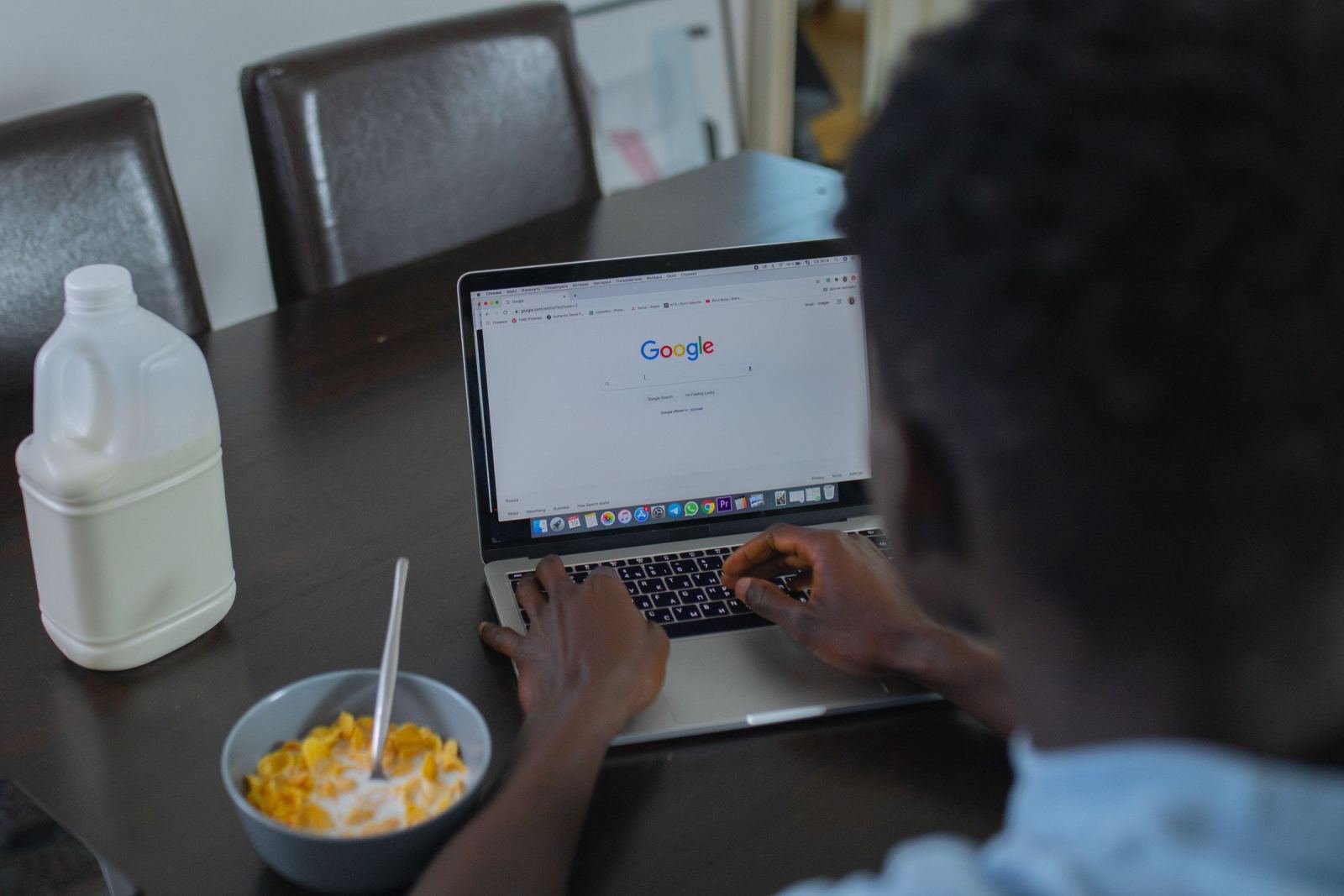 A person using a laptop at a kitchen table with a bowl of cereal and milk