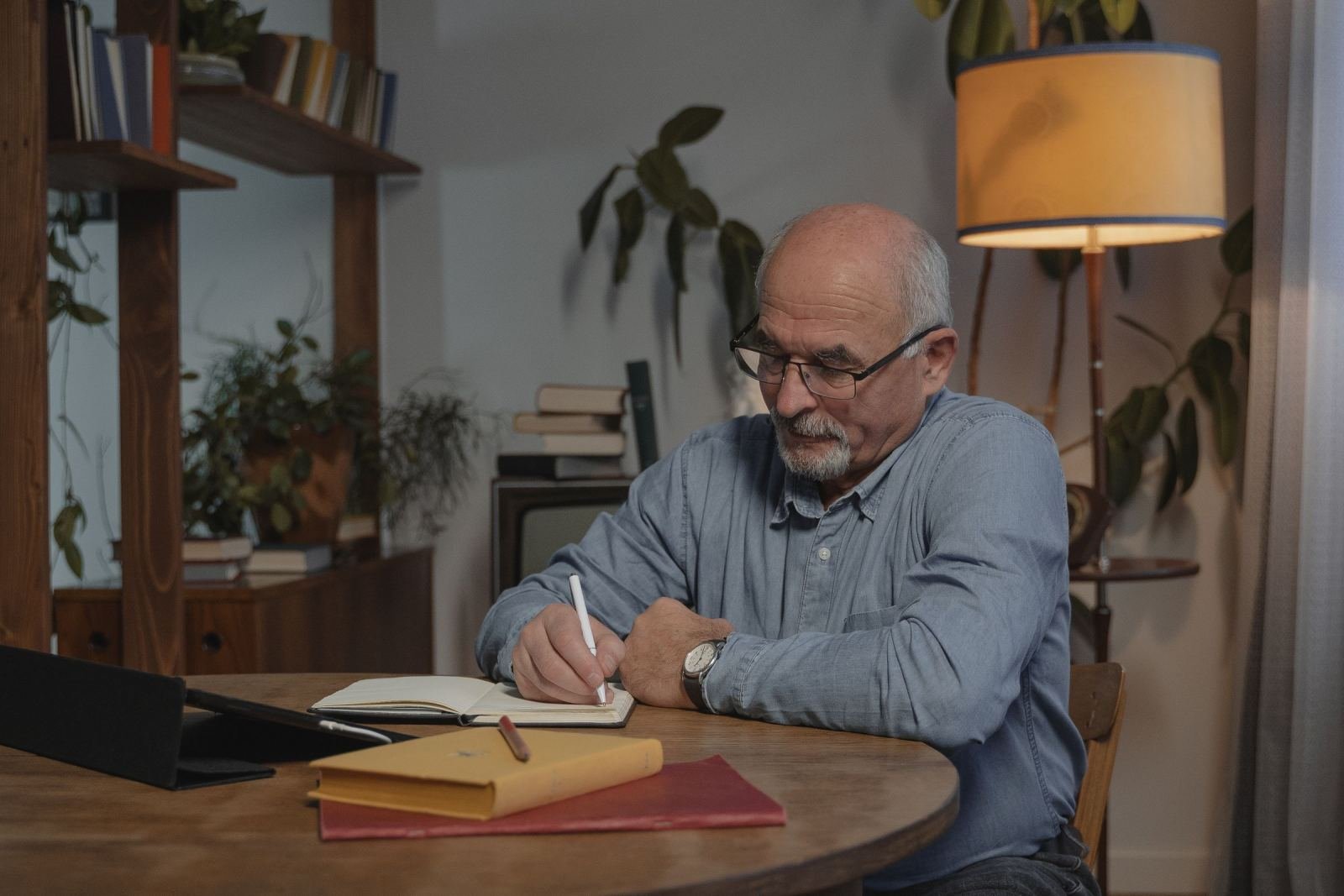 A senior man wearing eyeglasses is writing in a notebook at a wood table