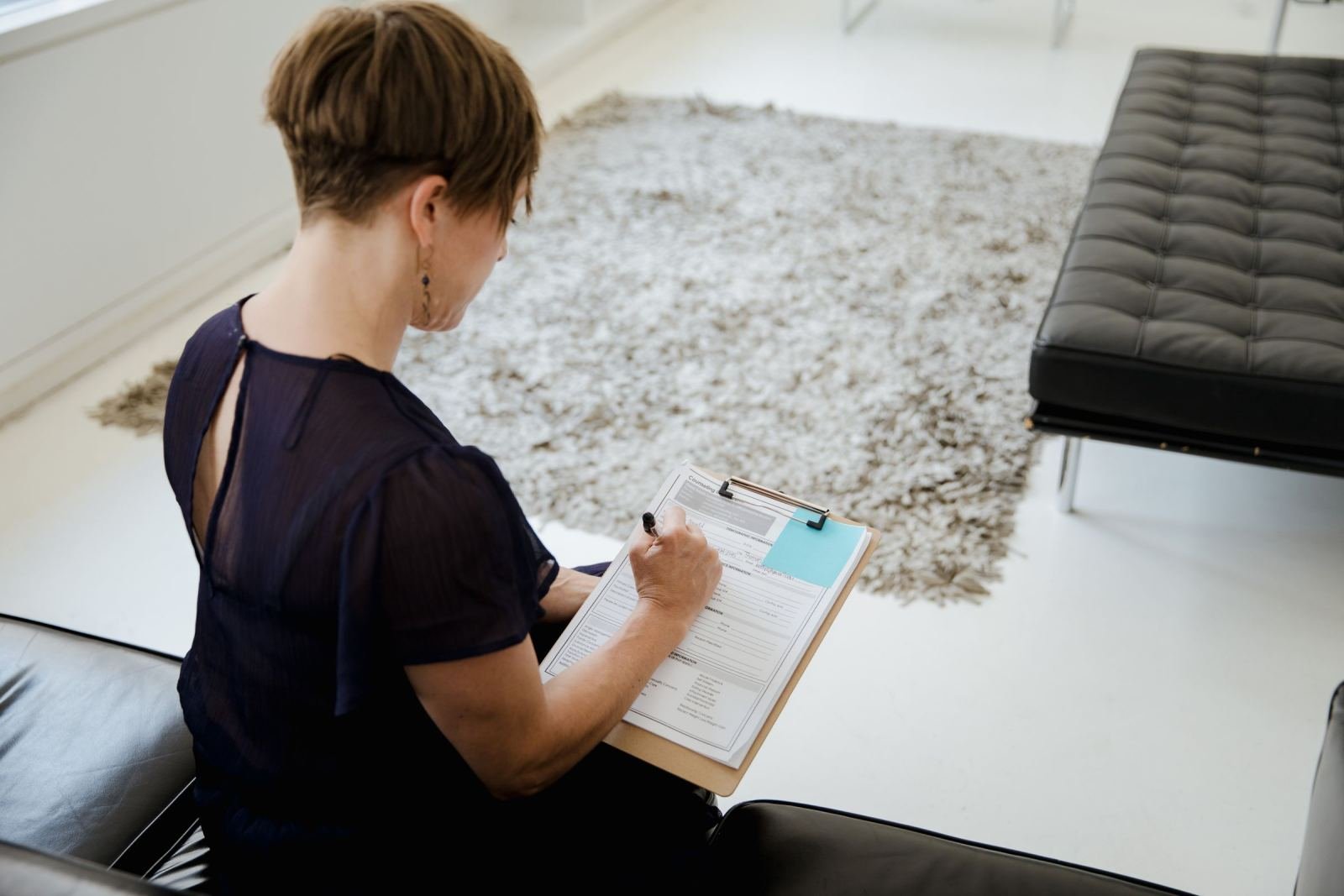 A woman sits on a black sofa and writes on a clipboard