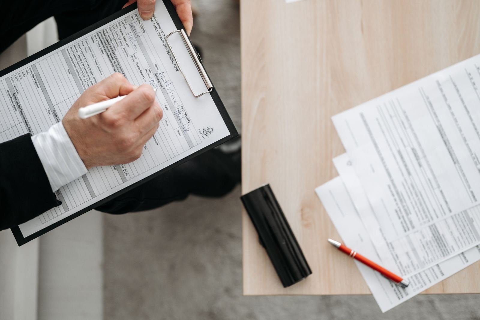 A person in a suit writes on a form on a clipboard at a wooden desk