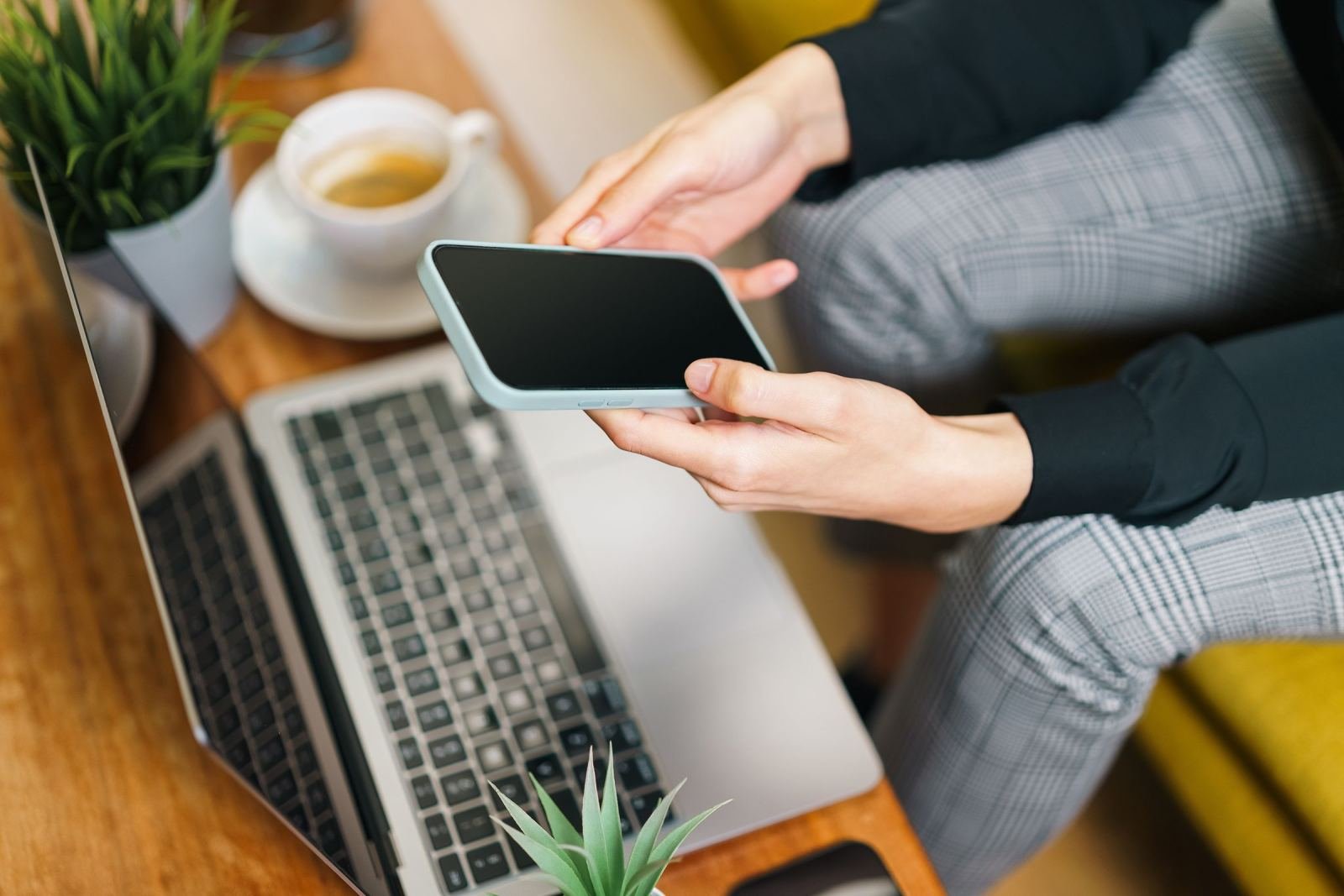 A person holding a smartphone while sitting at a table with a laptop and coffee