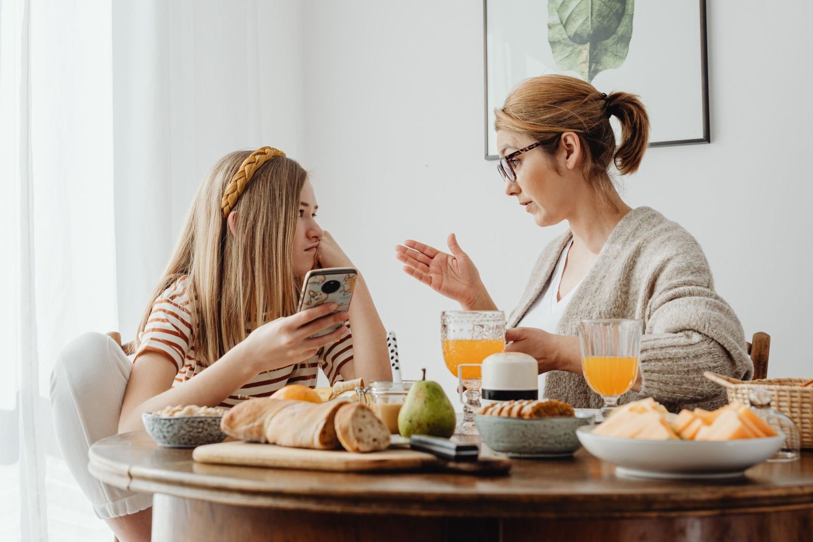 Two women sit at a kitchen table. They have breakfast and orange juice.