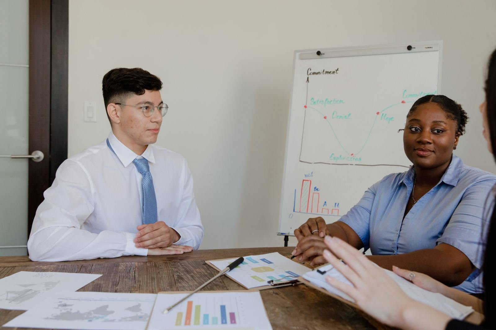 Two professionals sit at a table with charts and a whiteboard in a bright office