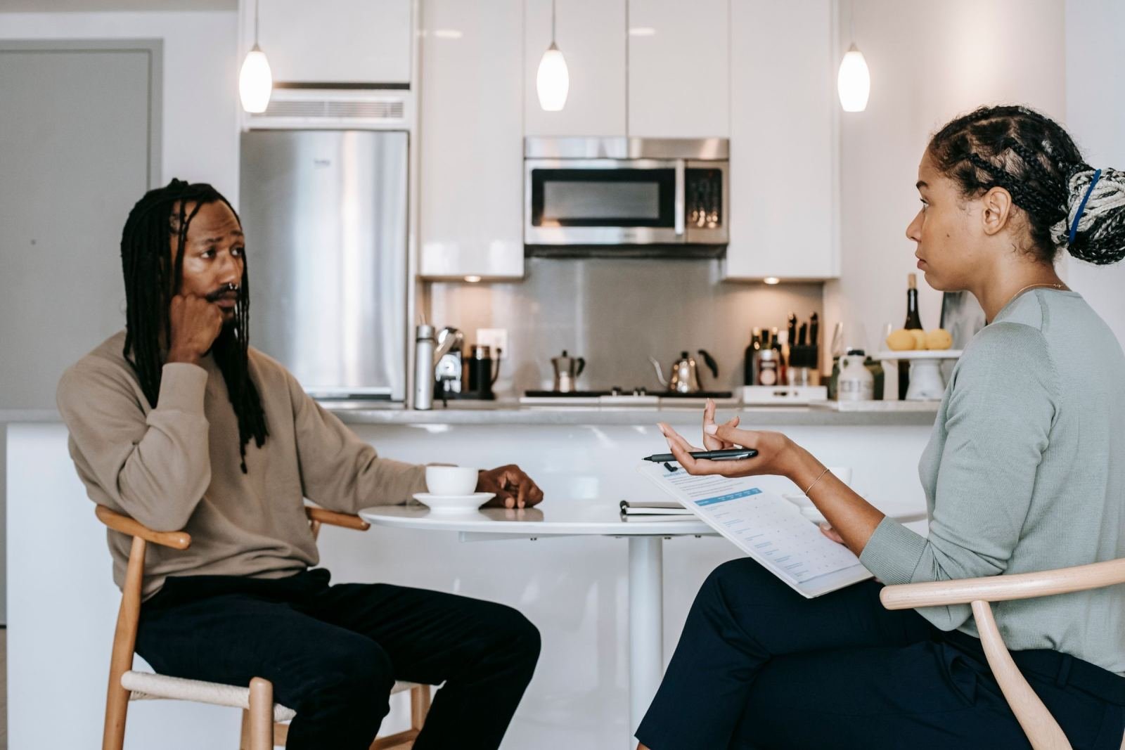 A man listens to a woman holding a clipboard while they sit at a white table