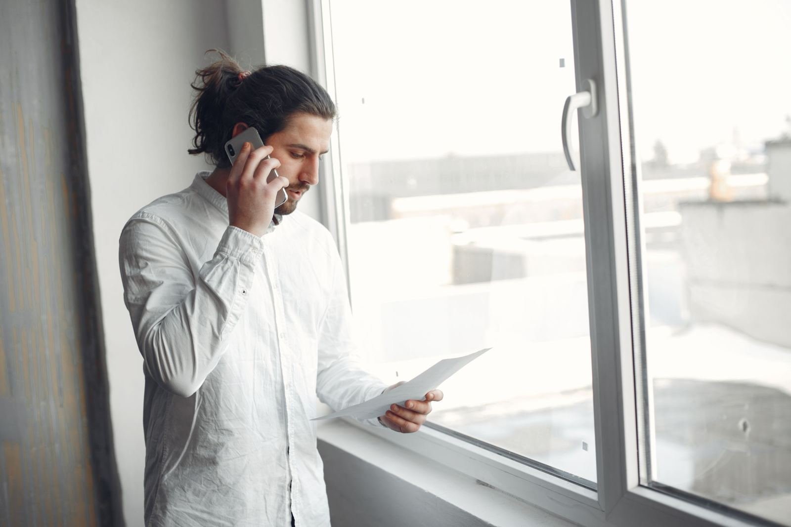 A man holds a mobile phone to his ear while he reads a paper near a window.