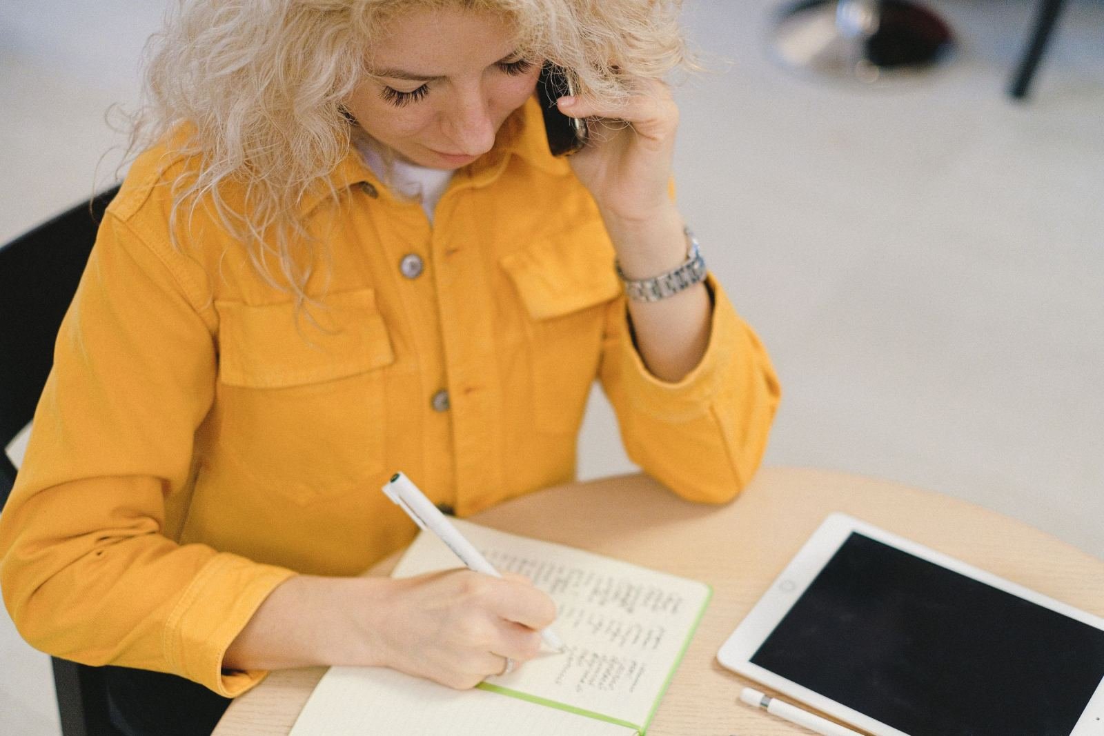 A woman in a yellow jacket talks on the phone while writing in a notebook