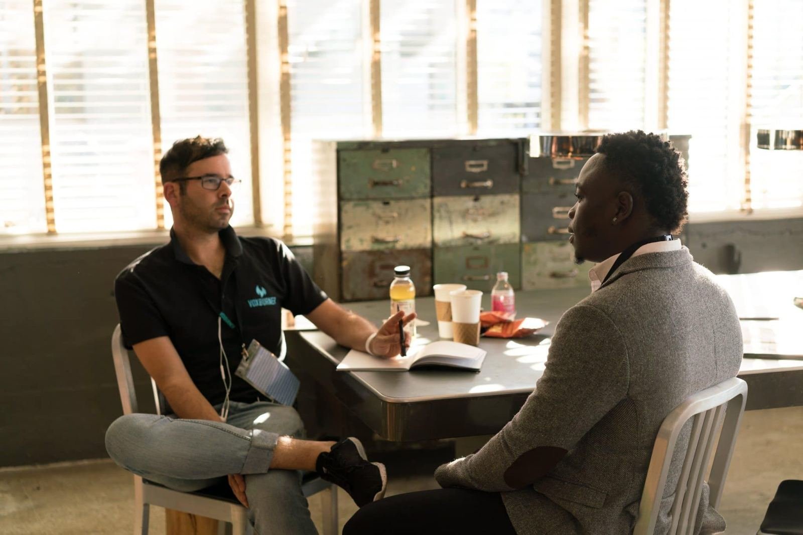 In a bright office, two men sit at a table reviewing a notebook together.