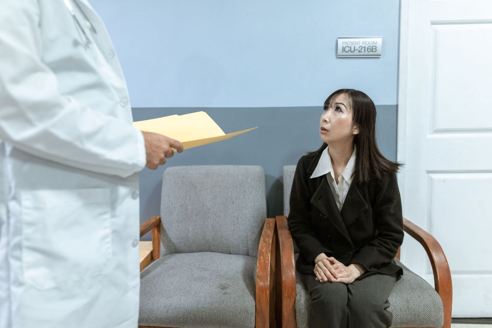 A clinician in a white coat holds a folder while talking to a woman sitting in a chair