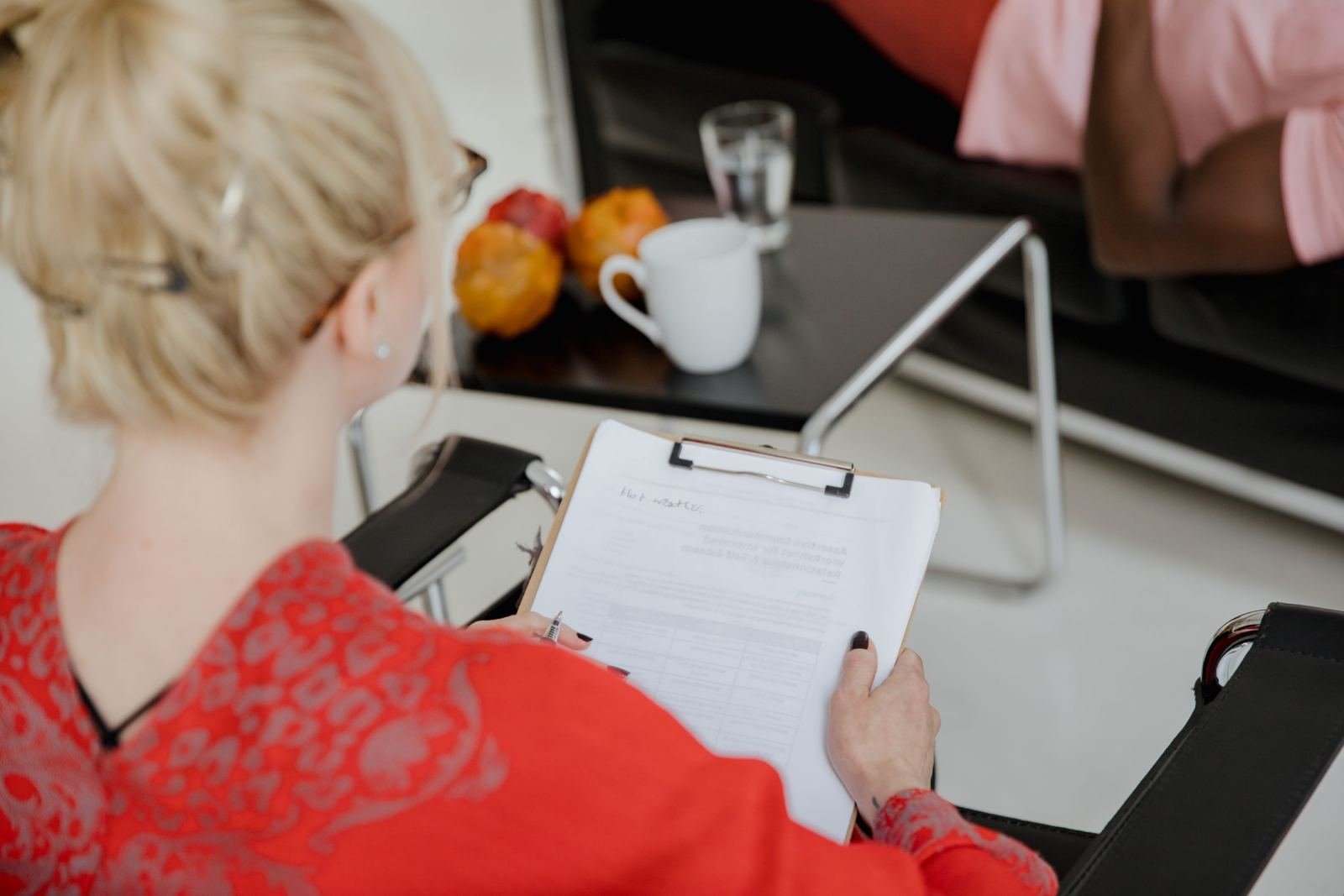 A person in a red shirt holds a clipboard while sitting across from someone in an office