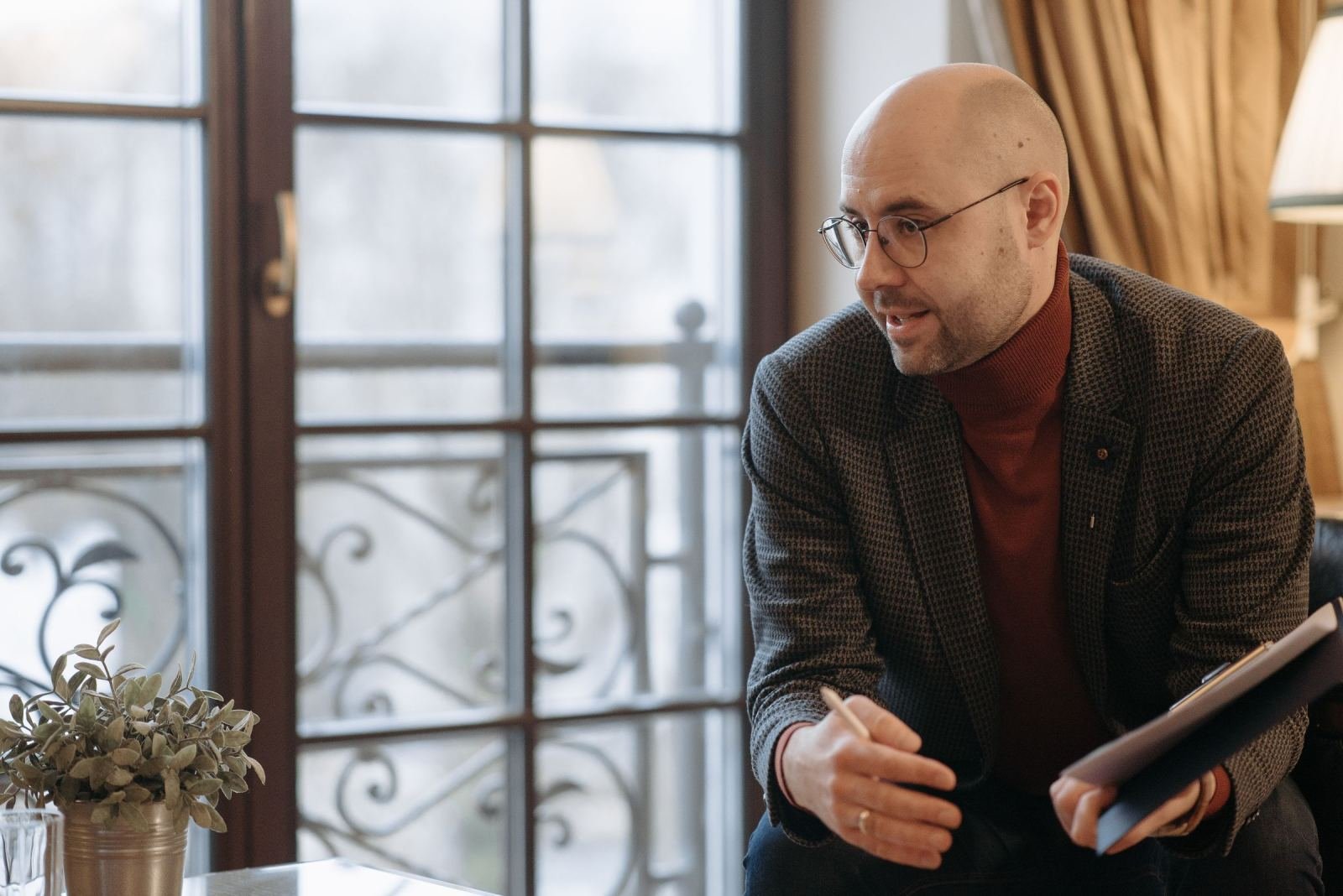 A man with glasses and a clipboard sits in an office and speaks to someone