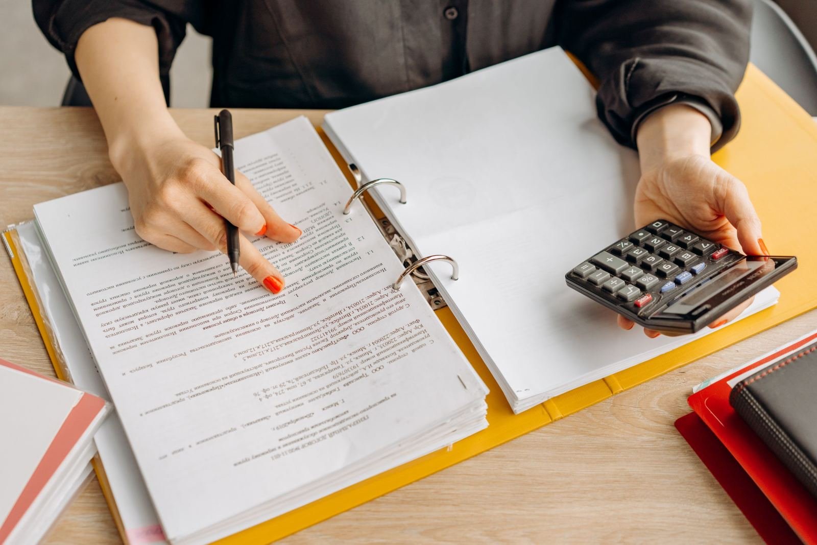 Someone holds a pen and calculator while checking papers in a folder.