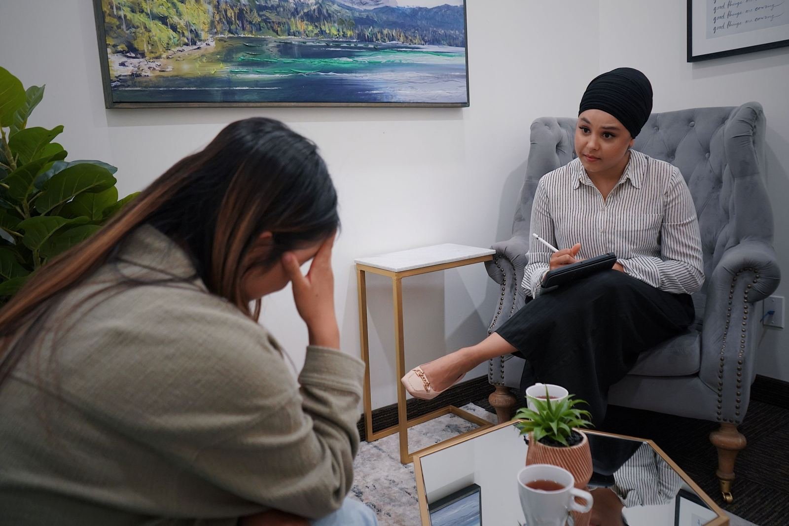 A woman sits in a grey armchair, hand on her forehead, while a clinician talks with her.