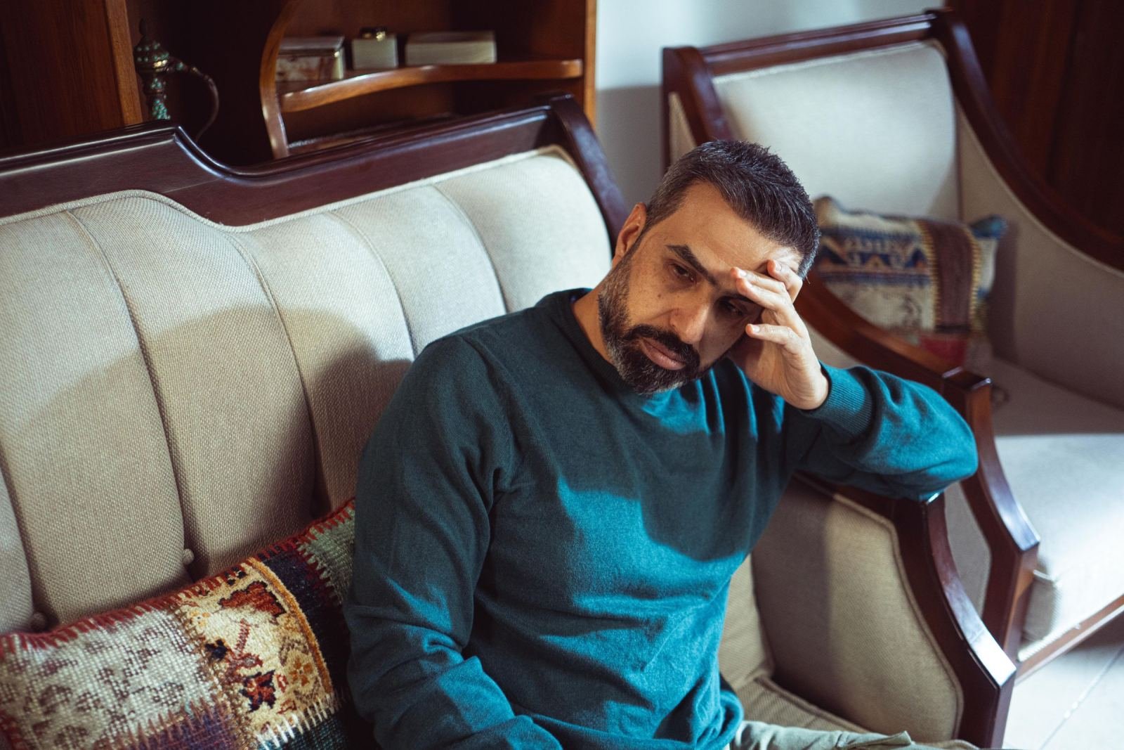 A man wearing a teal sweater sits on a couch with his hand on his head