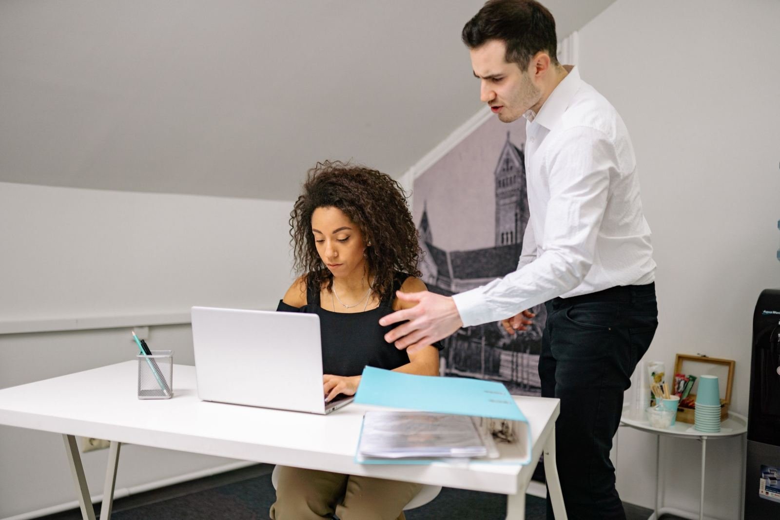 A man stands and gestures toward a laptop screen while a woman sits at a desk