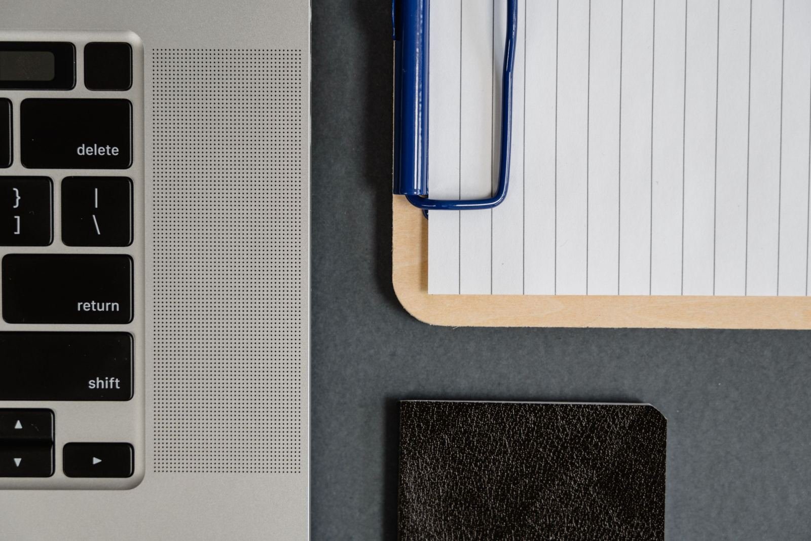 A laptop, a lined notepad on a clipboard, and a small black notebook on a dark desk