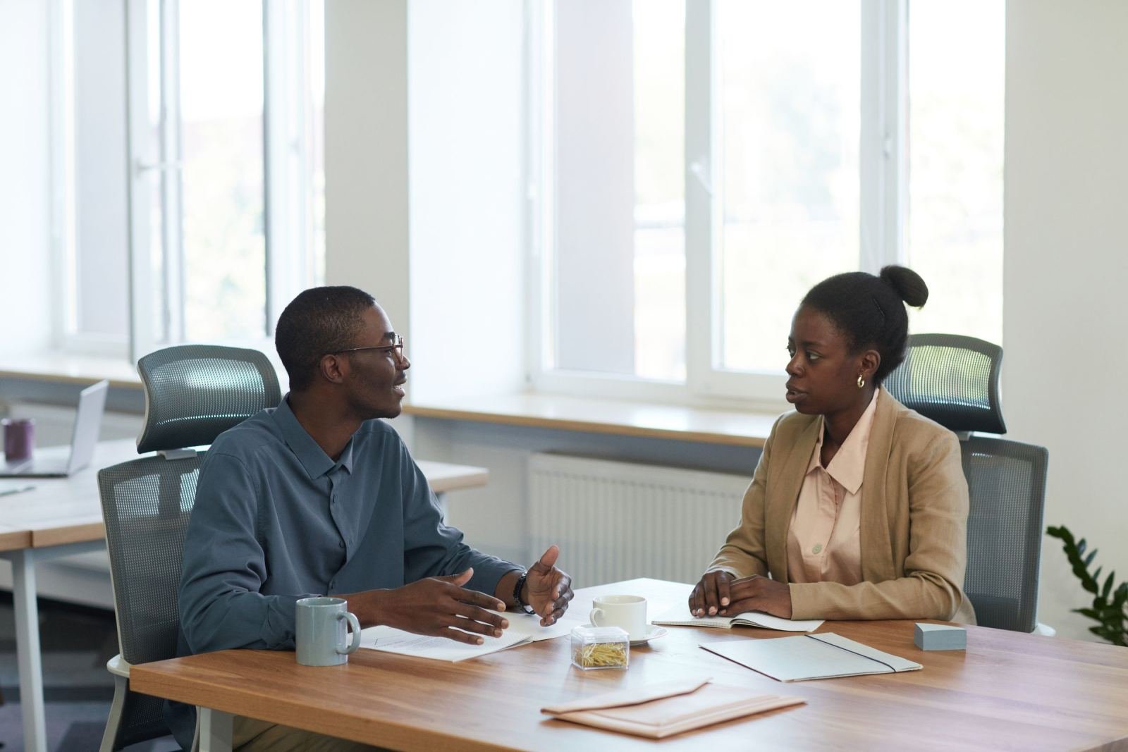 A man and woman talk across a wooden table in a bright office