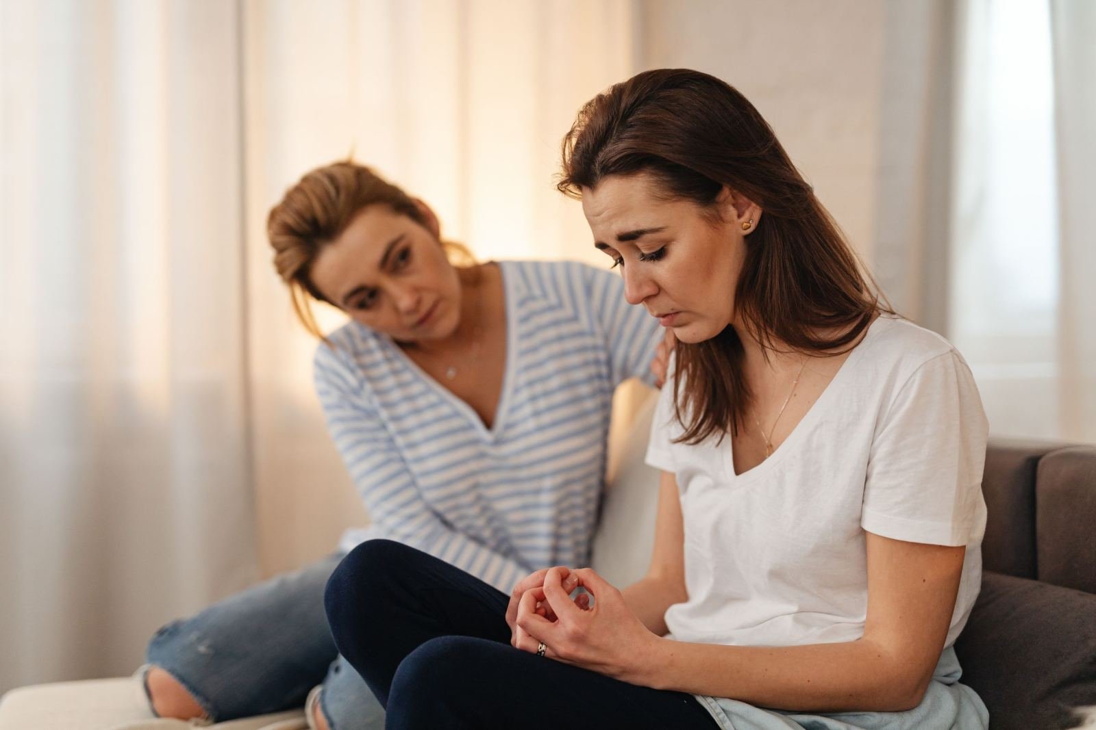 A woman wearing a striped shirt puts her hand on another woman's shoulder. The second woman is looking down.