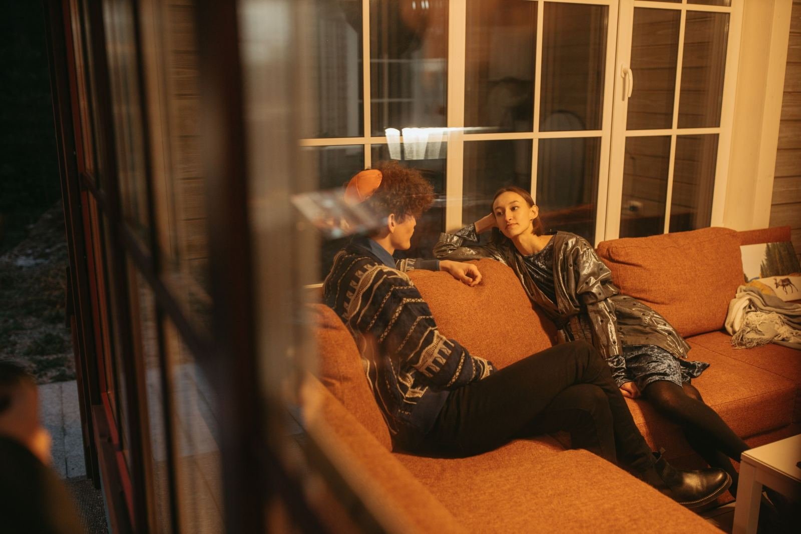 Two people sit on an orange sofa and talk in a room with large windows