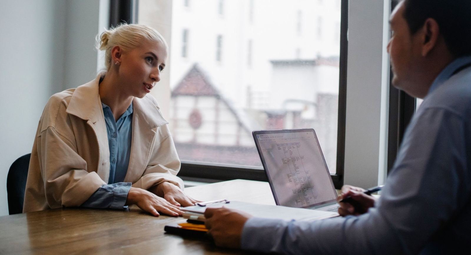 A man sits across a table from a woman in a tan jacket. A laptop rests on the table between them.