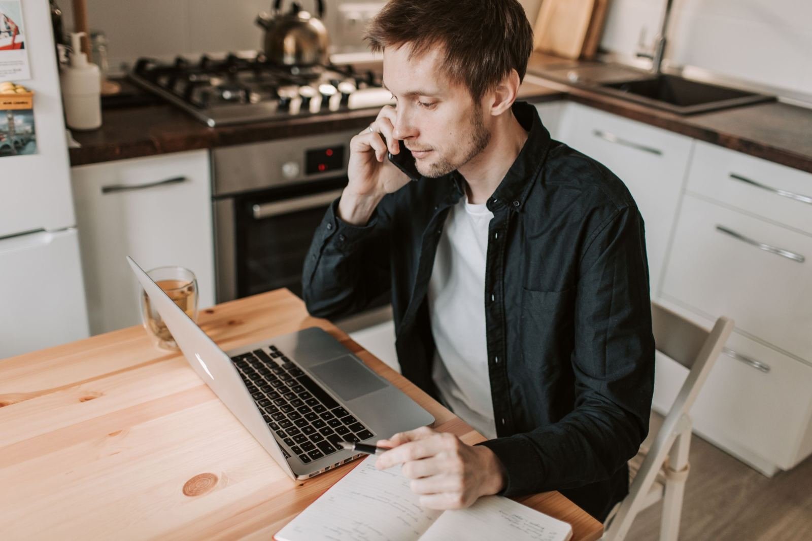 A man on a phone call sits at a kitchen table with a laptop and notebook