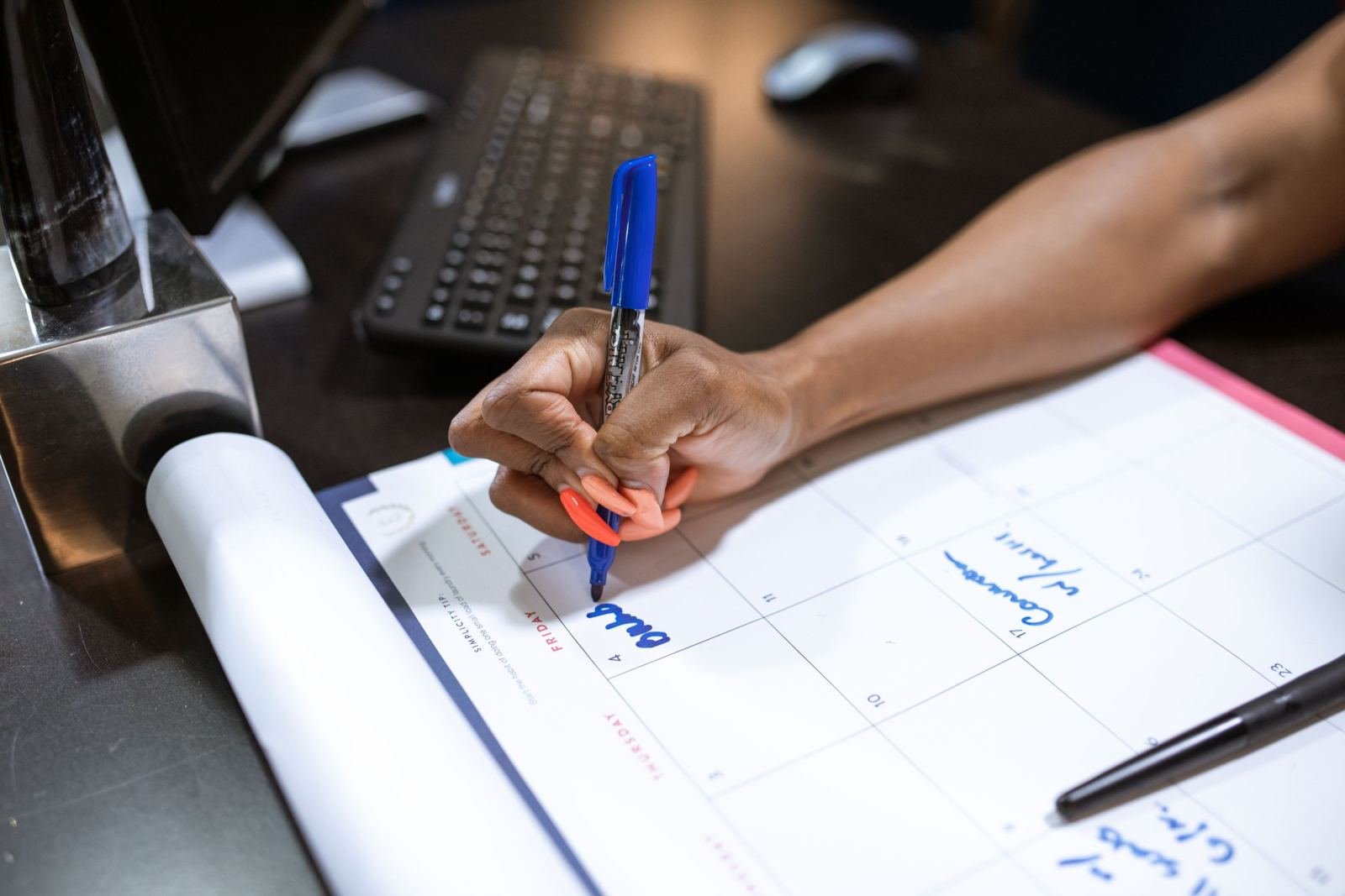 A person uses a blue pen to write on a weekly calendar at a desk