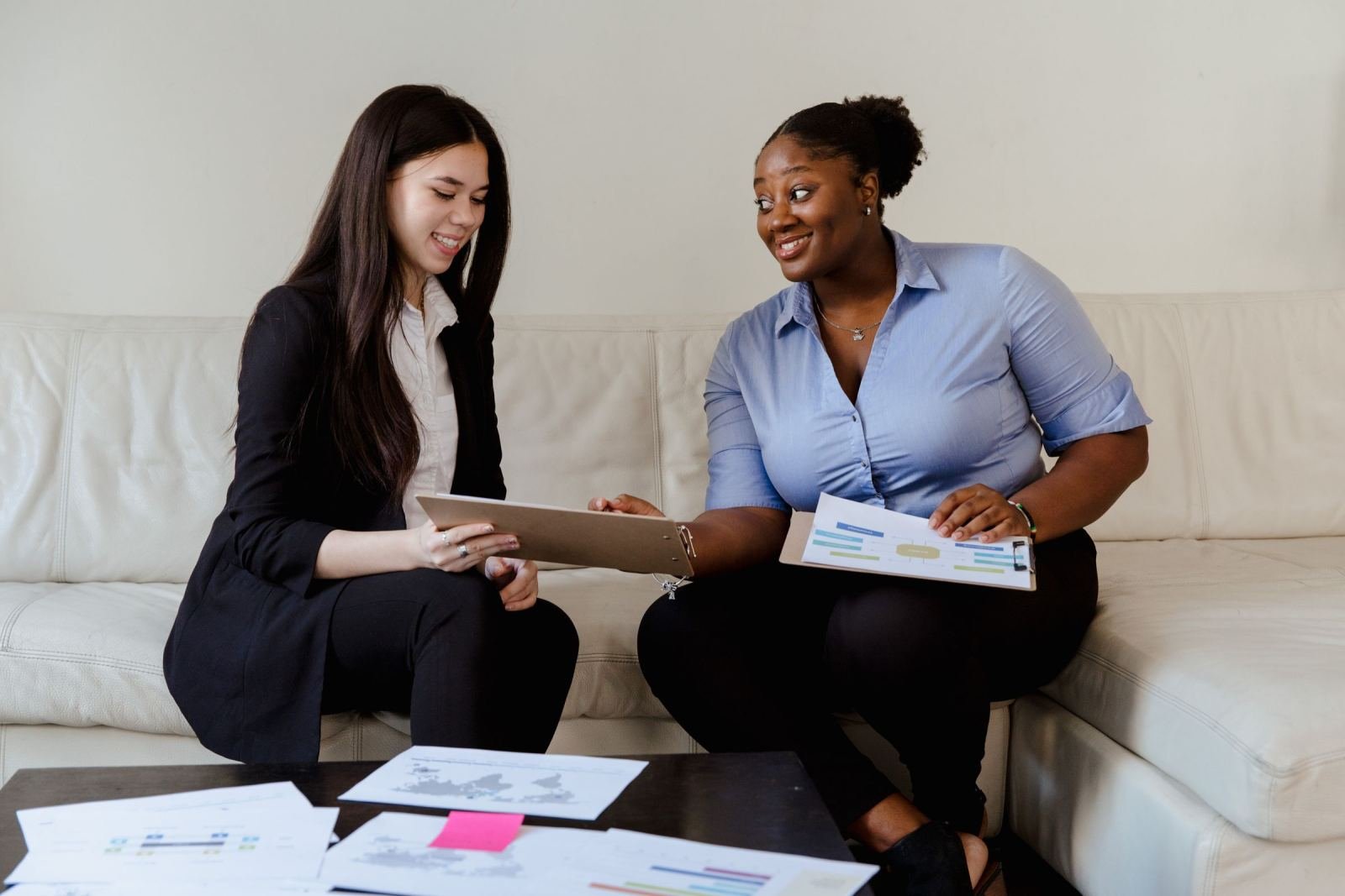 Two women sit on a white sofa reviewing documents and smiling