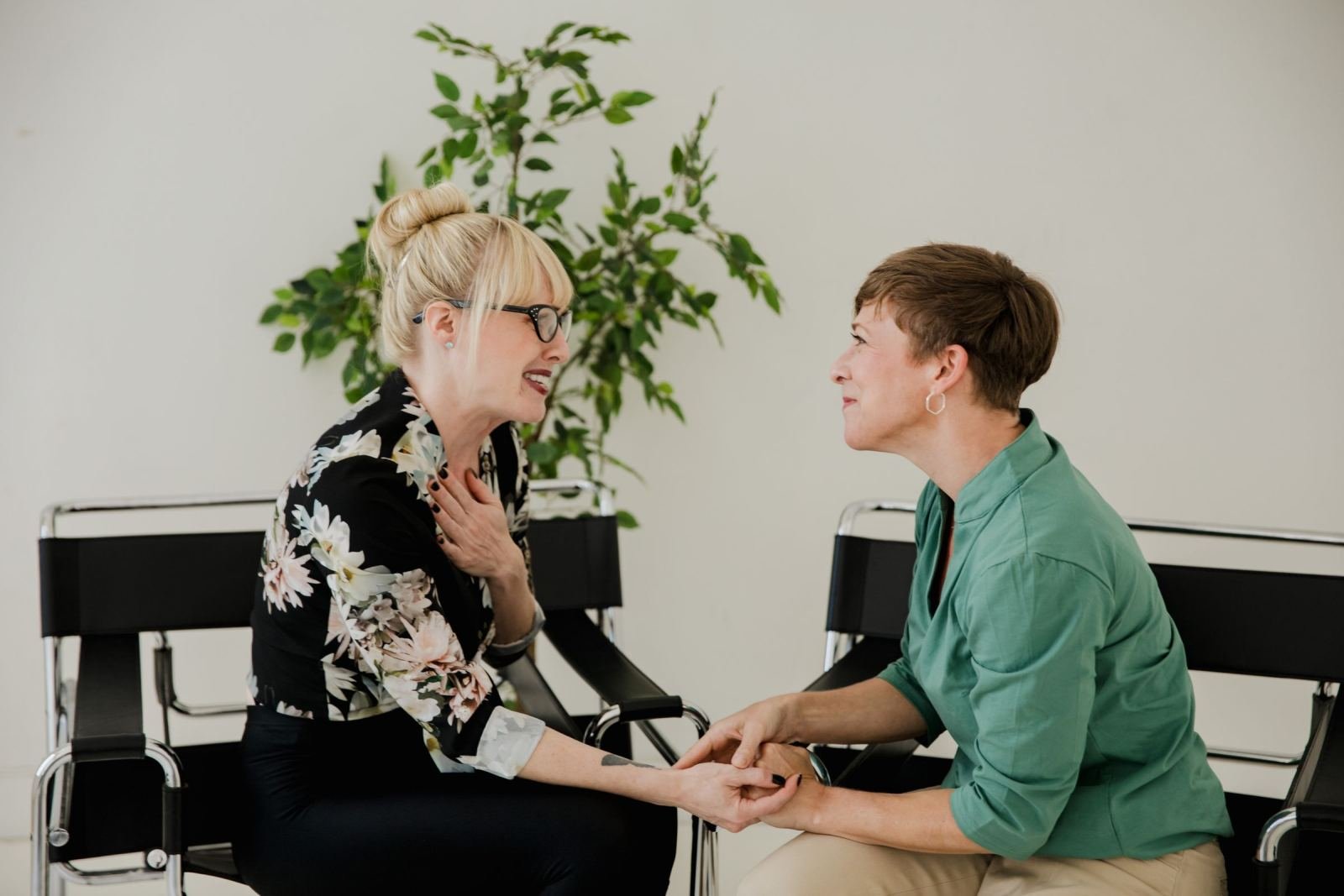 Two women sit in chairs facing each other, holding hands in a bright room