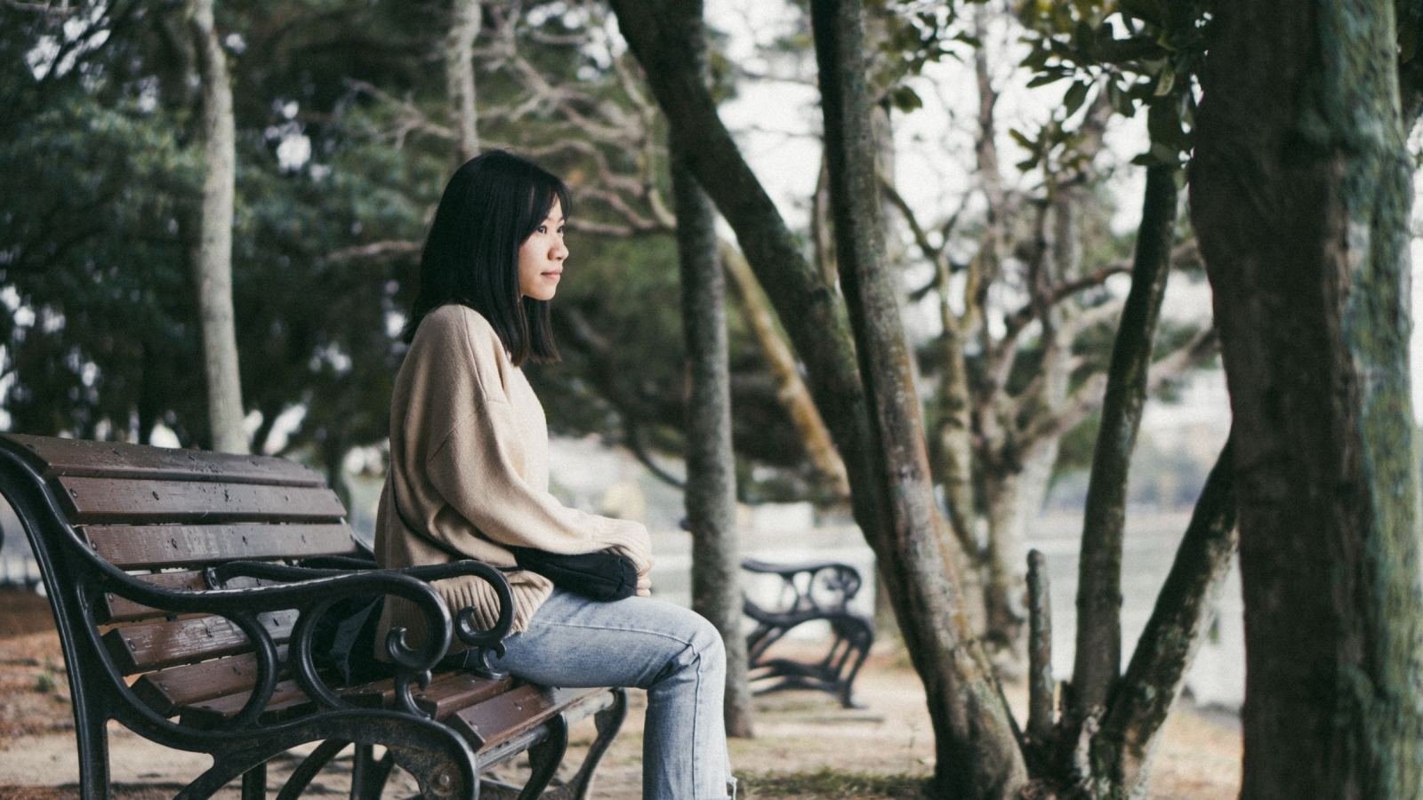 A woman in a beige sweater sits on a wooden park bench among trees