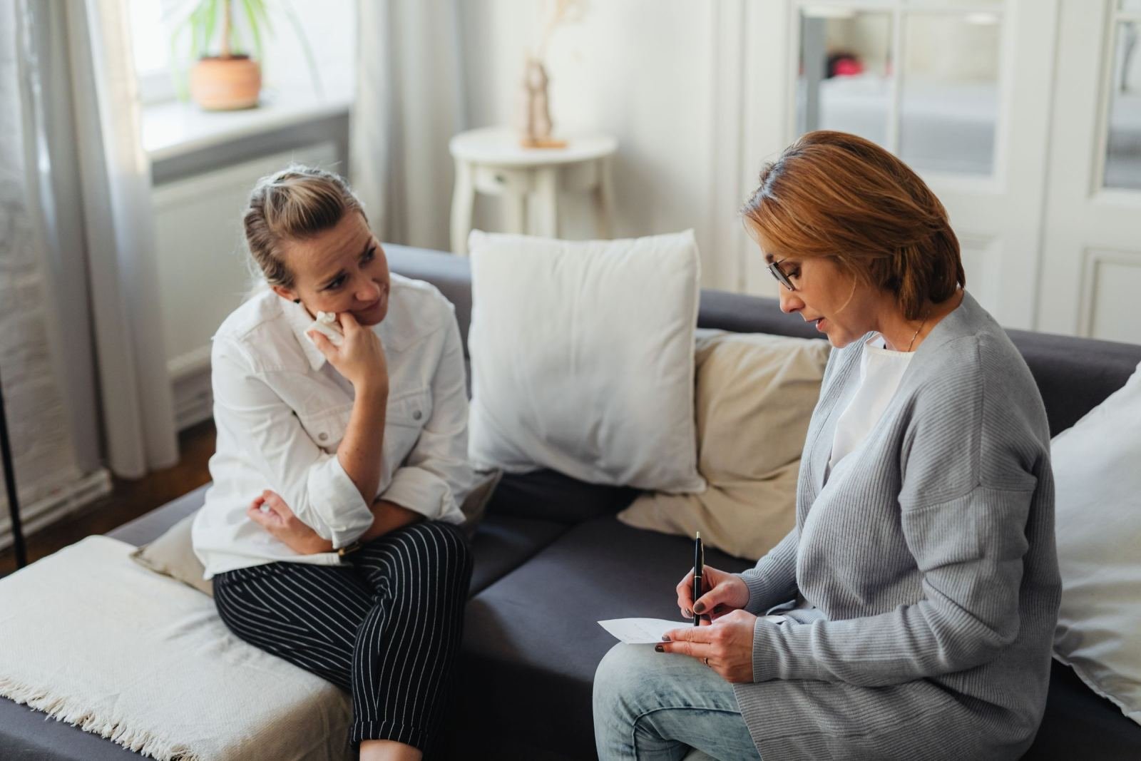 Two women sit on a sofa talking while one takes notes on a piece of paper