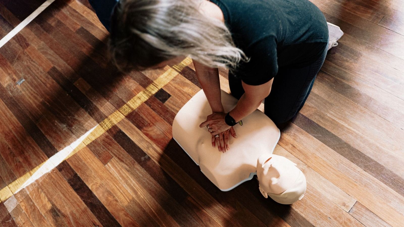 Someone does chest compressions on a CPR dummy that sits on a wood floor.