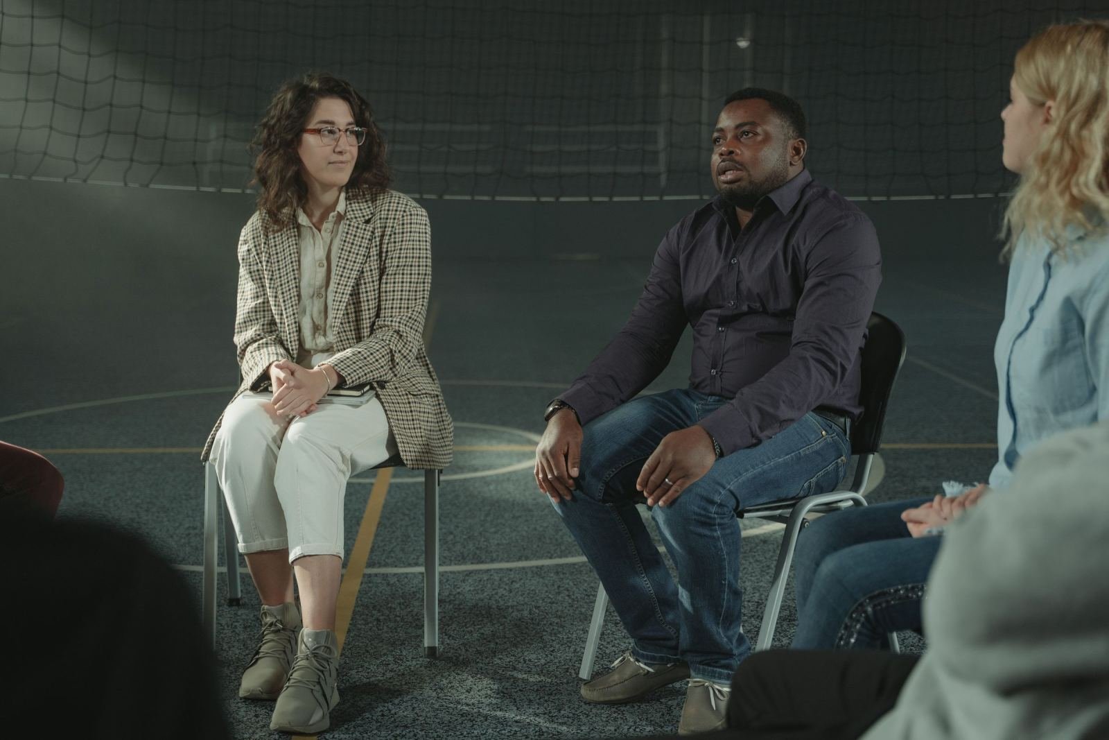 Three adults sit in a circle of chairs and talk in a dimly lit room