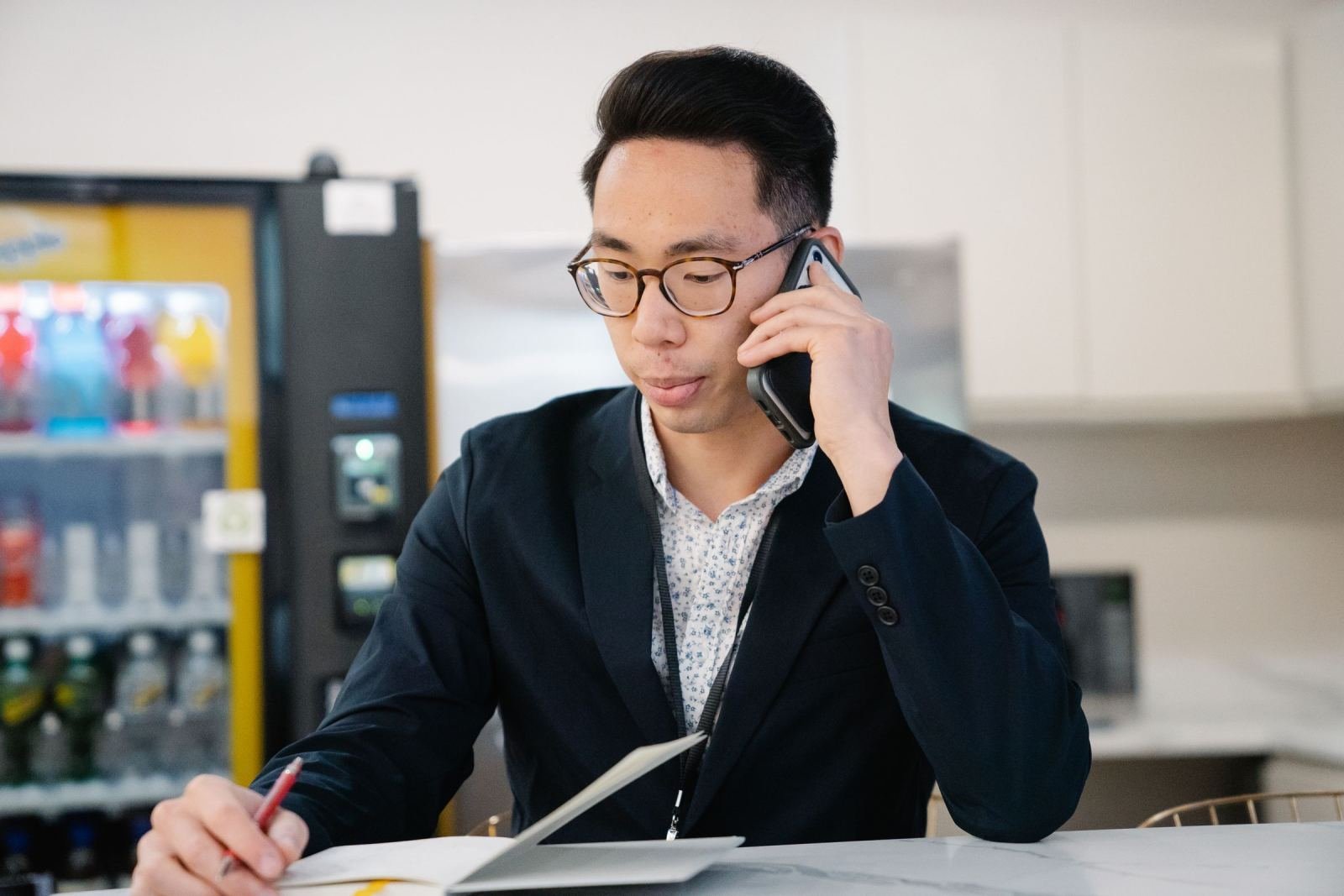 A man in a blazer talks on a phone while writing in a notebook