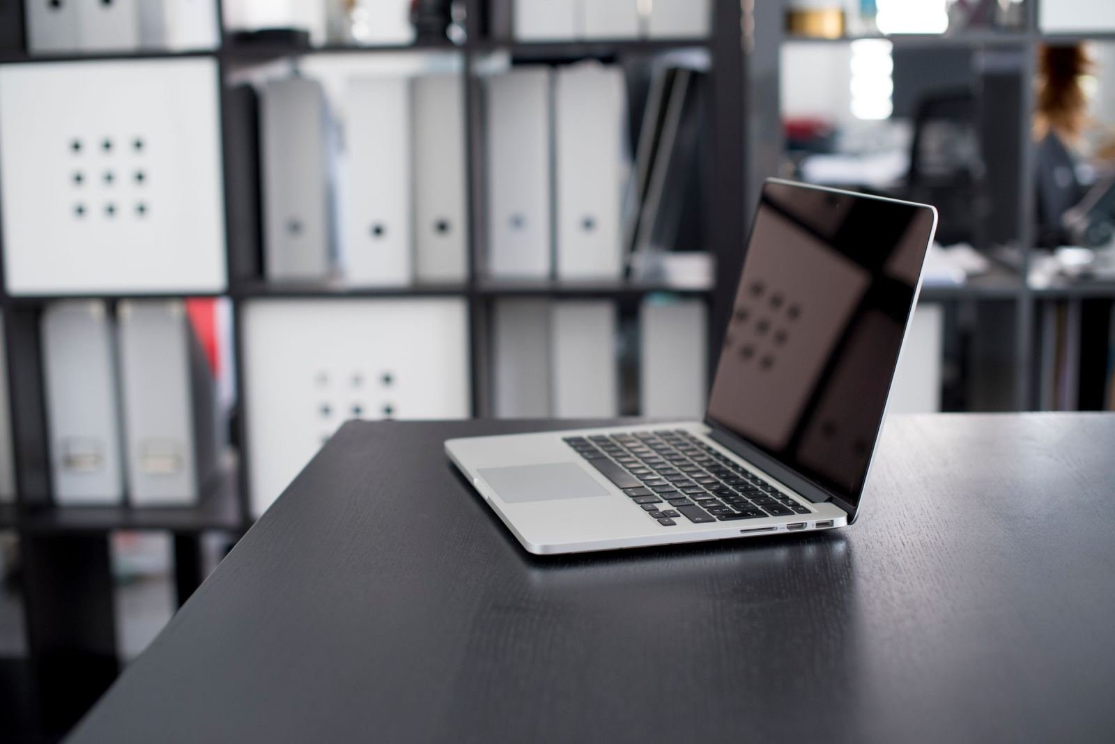 A laptop sits open on a dark desk. Blurred office shelves sit behind it.