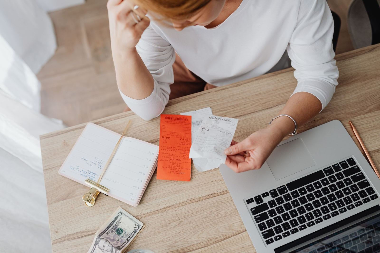 A person sorts receipts and bills at a desk with a laptop and cash