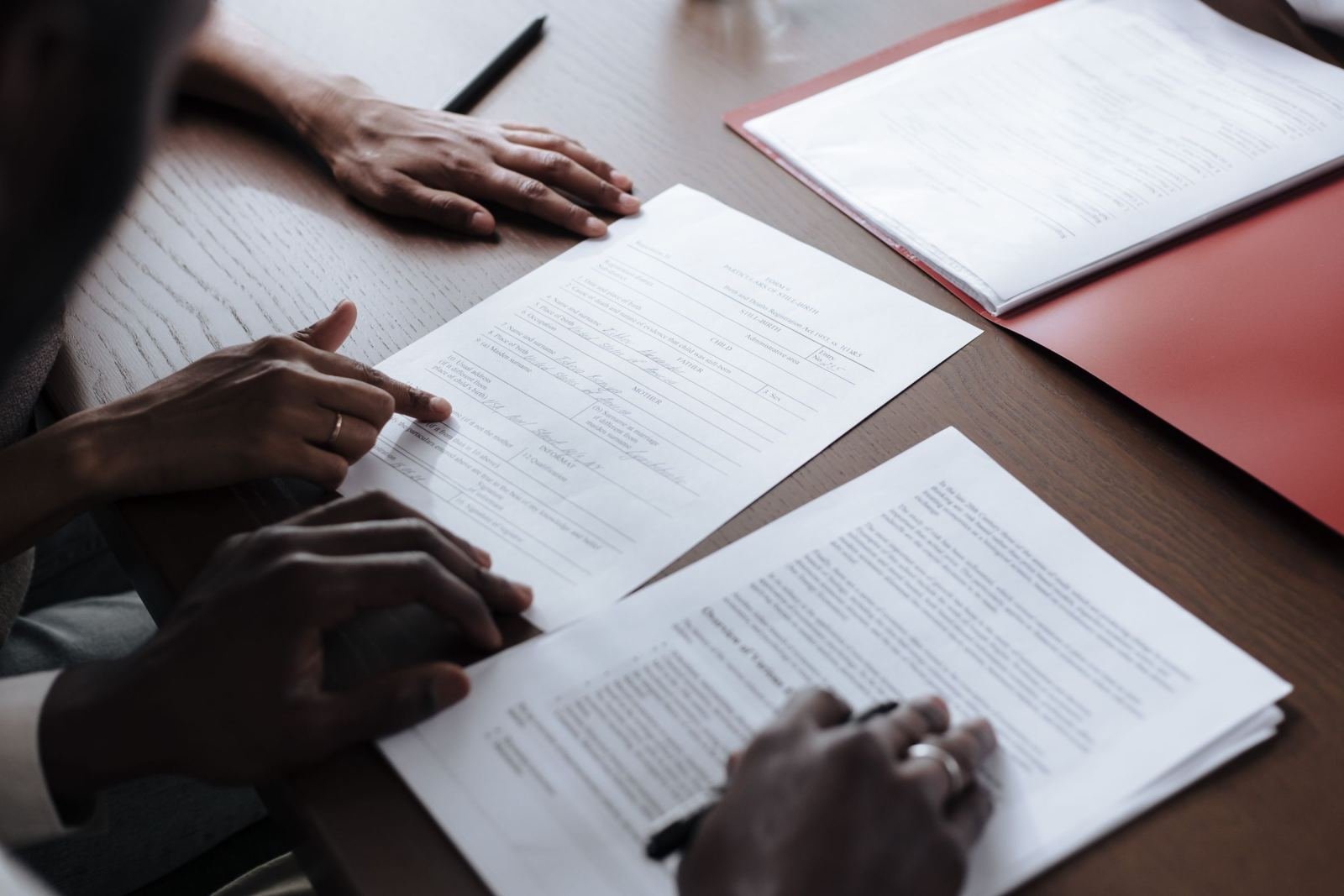 Two people sit at a wood table. They look at papers and point out parts of them.