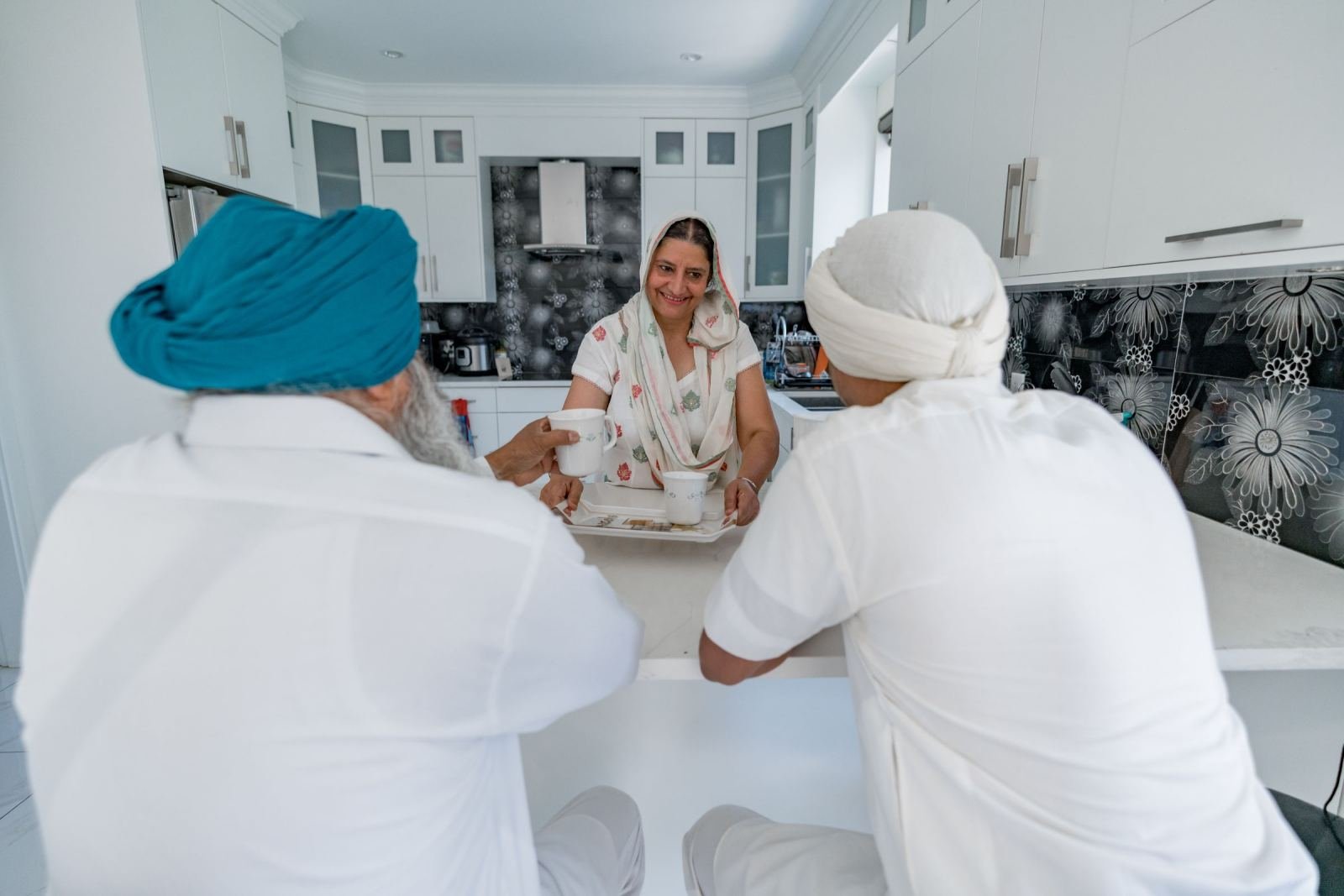 A smiling woman holds a tray of mugs while two men in turbans sit at a kitchen counter.