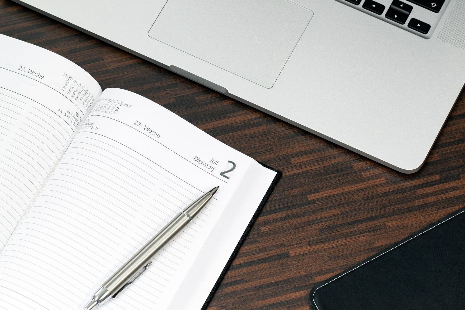 An open planner and a silver pen on a wooden desk next to a laptop