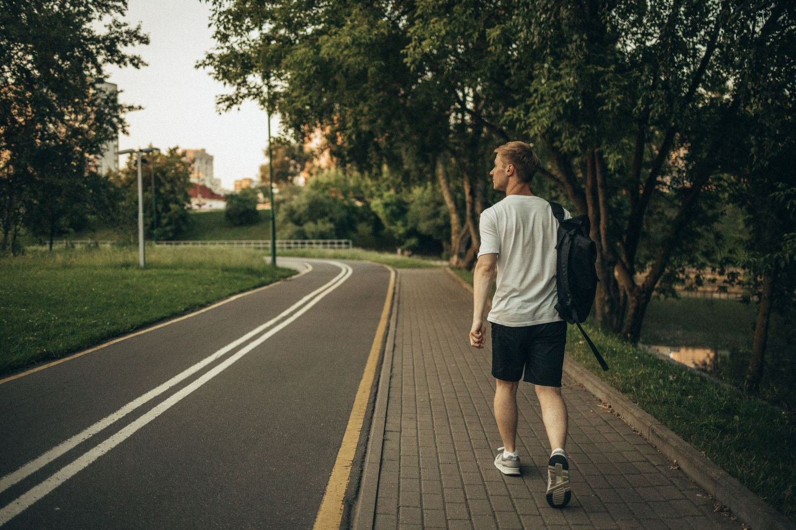 A man carrying a backpack strolls down a paved trail in a green park.