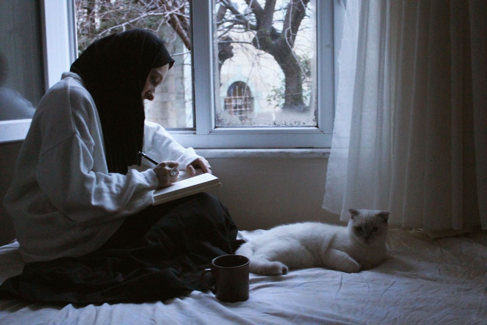 A person in a hijab writes in a journal on a bed next to a white cat