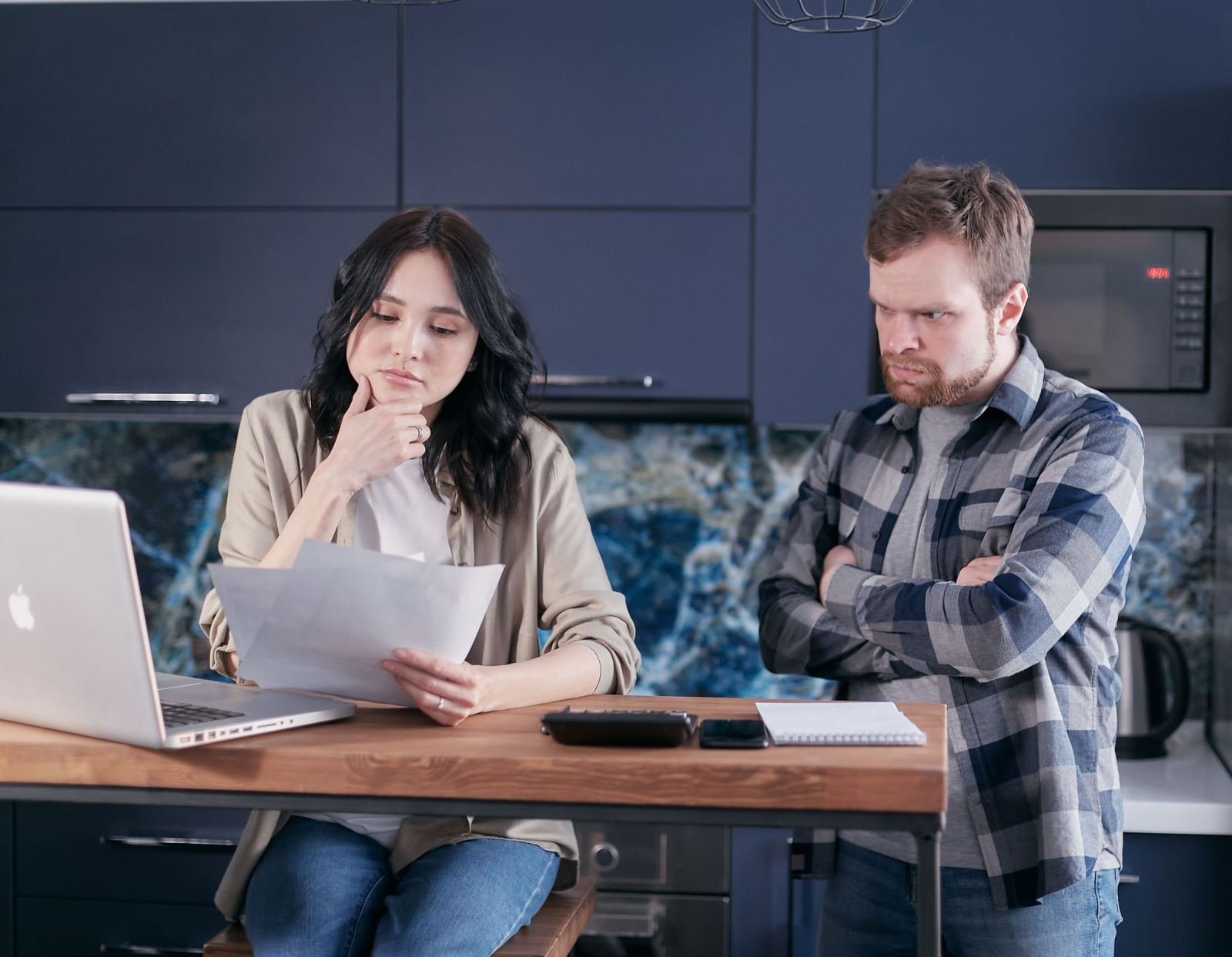 A woman reviews papers and a laptop while a man stands beside her with arms crossed