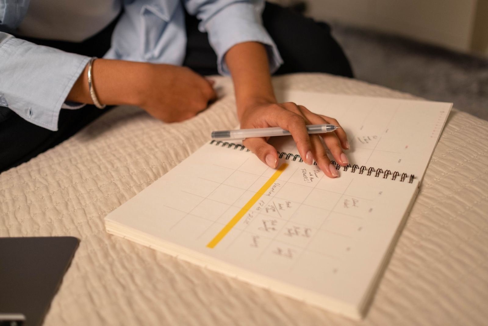 A person in a blue shirt writes a list in a spiral notebook on a white surface
