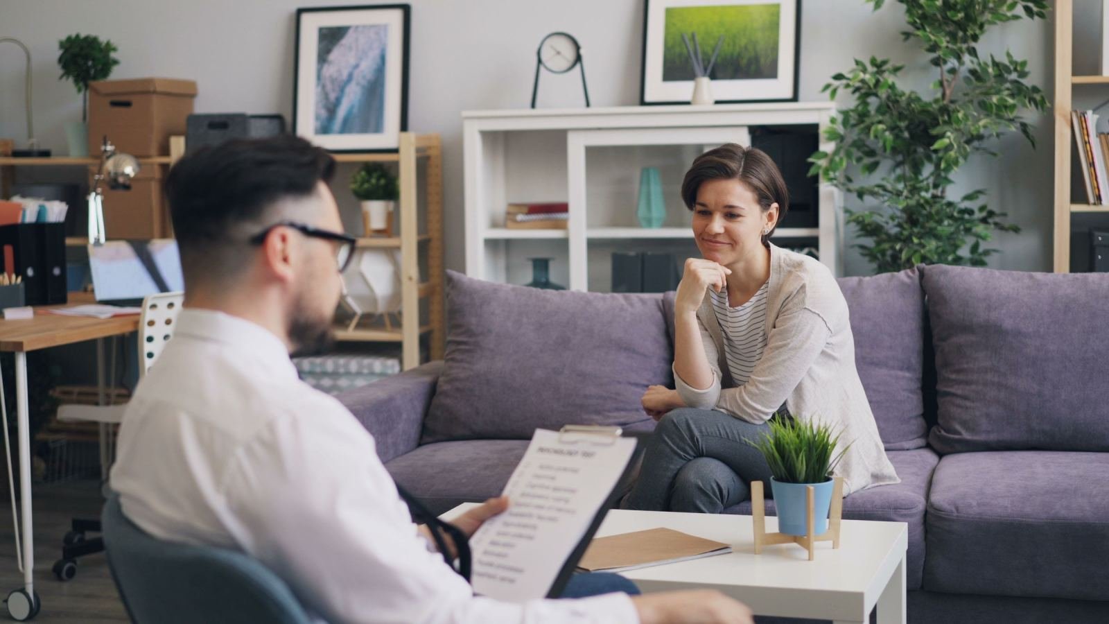 A counselor holding a clipboard speaks with a woman seated on a purple couch.