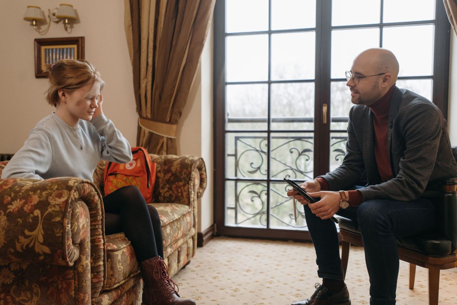 A woman sits on a sofa while a man with a tablet speaks with her in a room