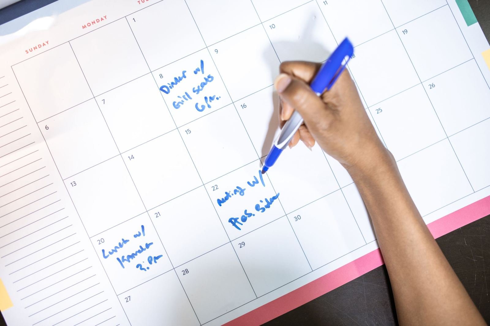 A hand uses a blue pen to mark an appointment on a paper calendar