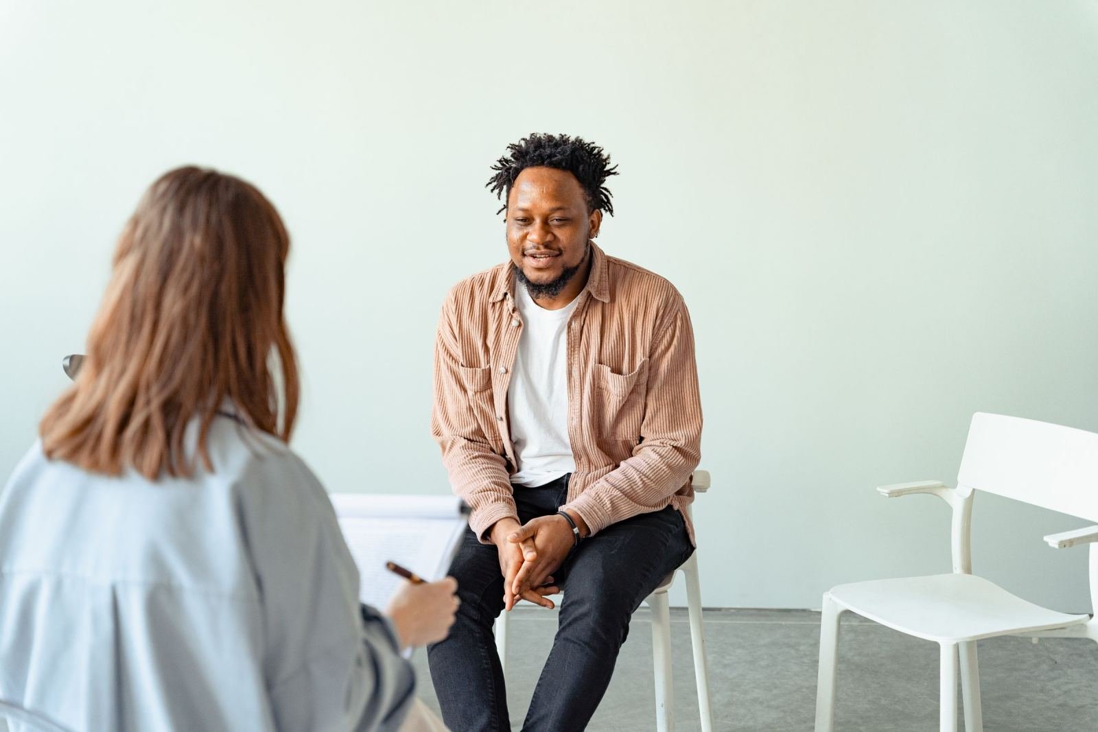 In a bright room, a man sits in a white chair while speaking with a counselor.