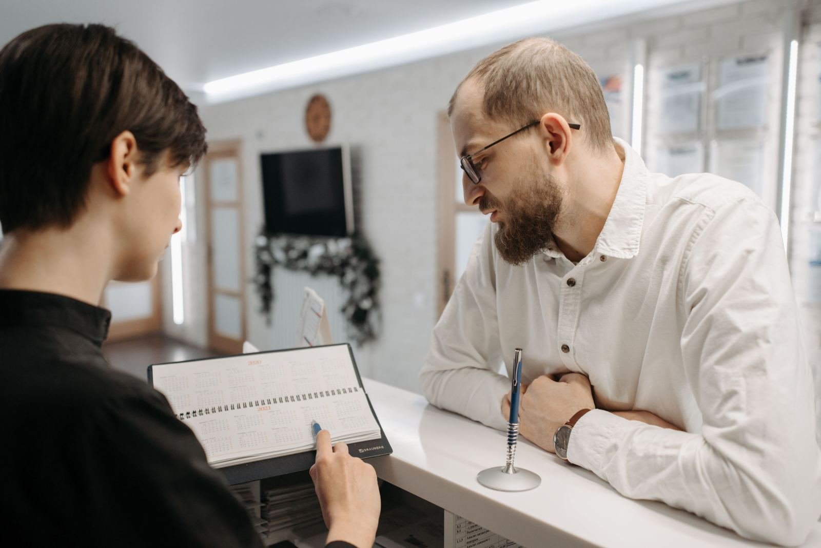A man speaks with a receptionist who is pointing to a planner on a white counter